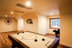 A room with a foosball table, a bear decoration, a compass wall decoration, and a window with a view of trees. - Luxury Cabin on Golf Course / Private Spa & Launch (Lake Almanor West)