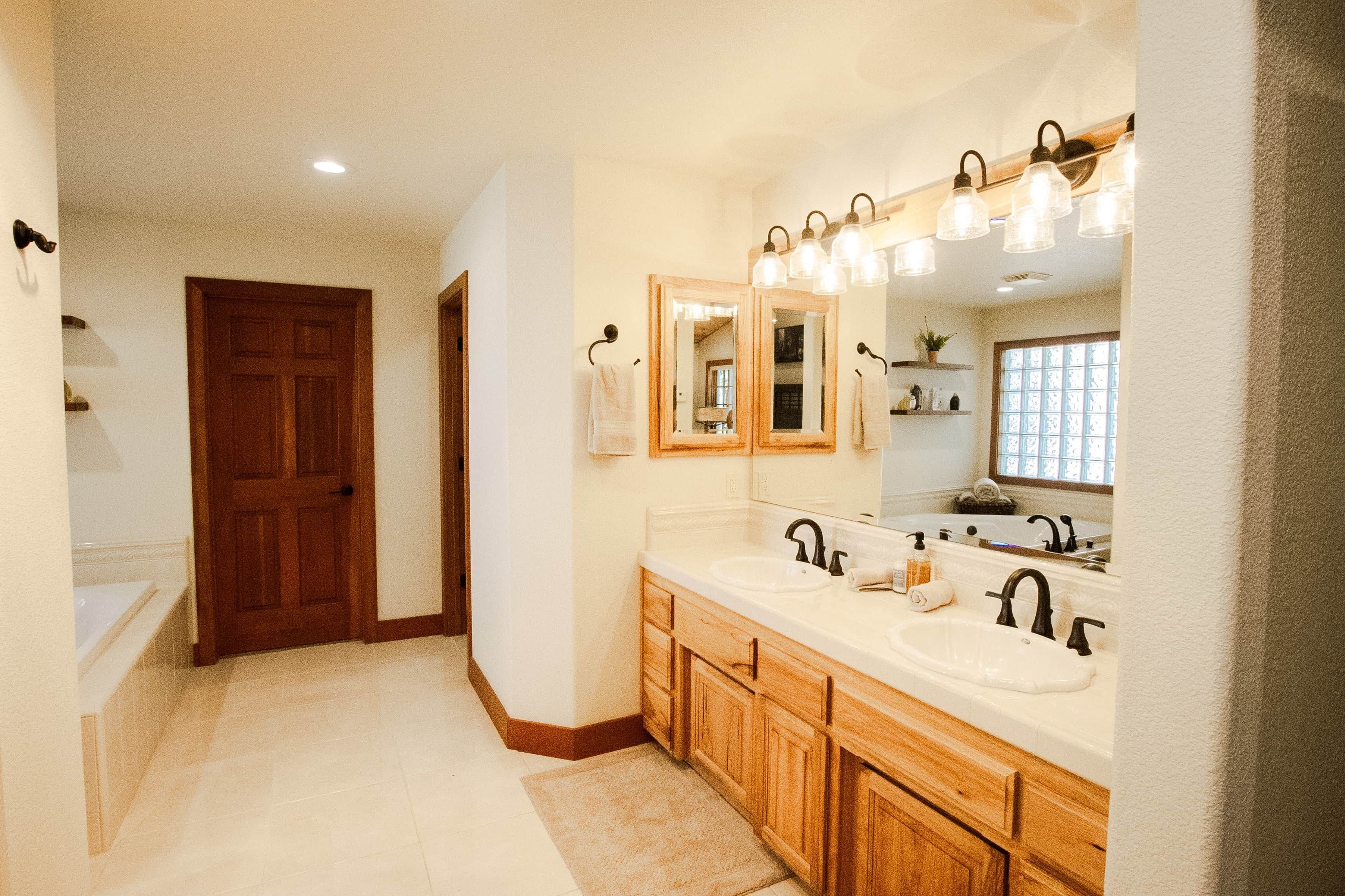 Une salle de bain avec un double lavabo, des meubles en bois, un grand miroir et une porte donnant sur une autre pièce.