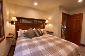 A bedroom with a wooden headboard, two bedside lamps, a bed with a plaid bedspread, and a wooden door.