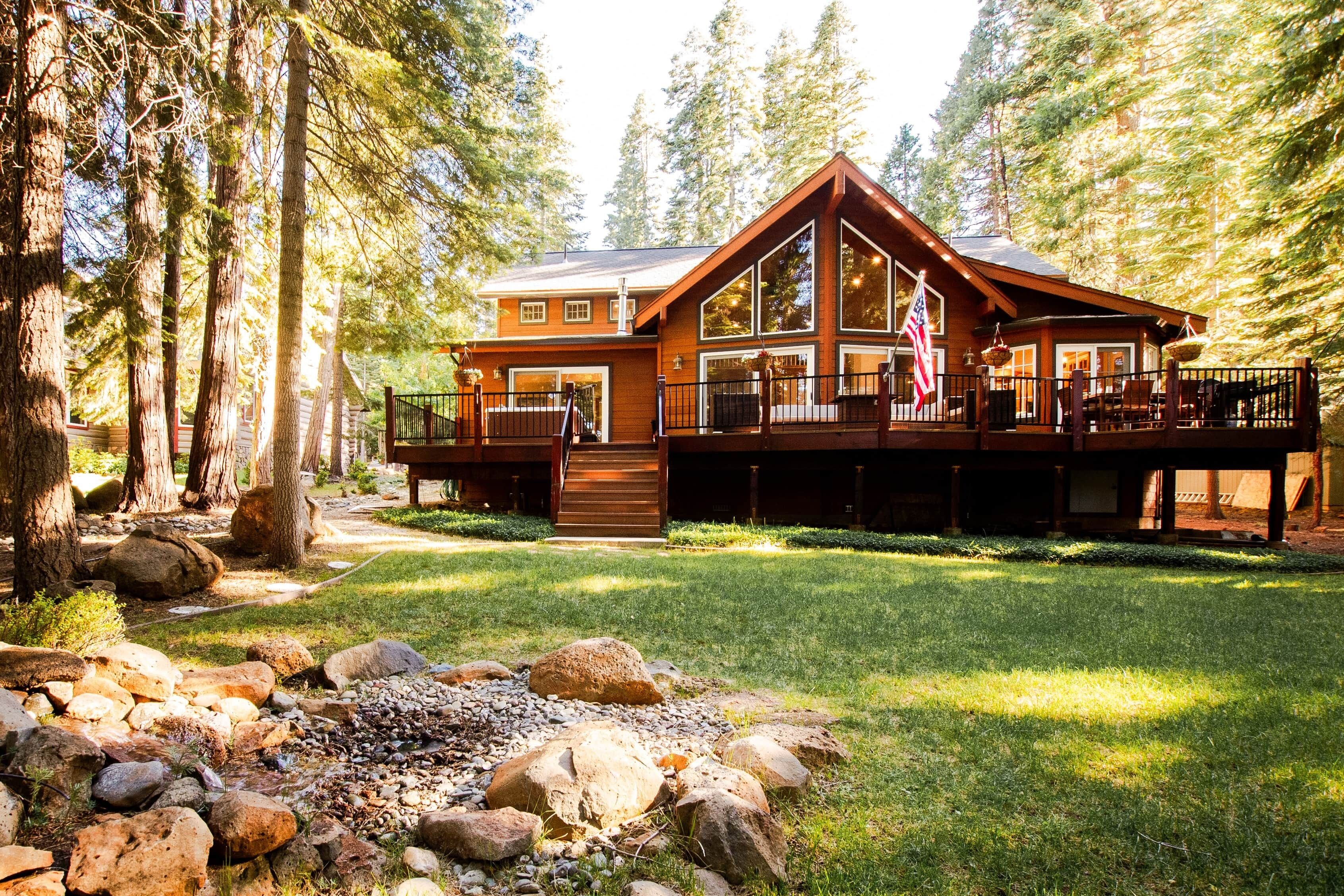 A cabin with a deck, surrounded by trees and a grassy area with rocks.