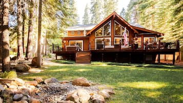 A cabin with a deck, surrounded by trees and a grassy area with rocks.