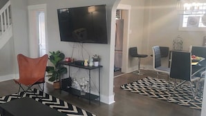 A living room with a staircase, a flat-screen TV, a dining area with chairs, and a patterned rug. - Perfect 3-bedroom house in the heart of Mt. Washingston (Pittsburgh)