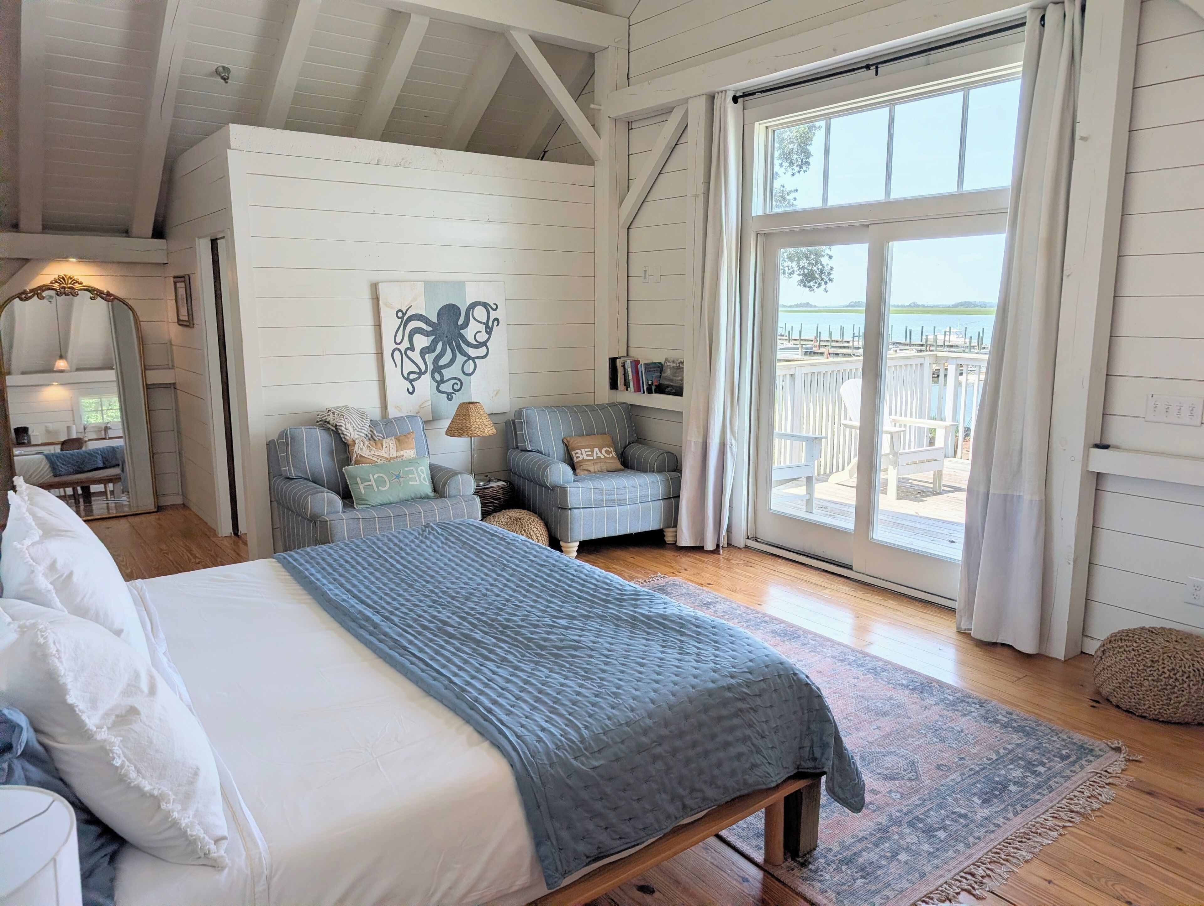 A bedroom with a large bed, two armchairs, a mirror, and a view of a dock.