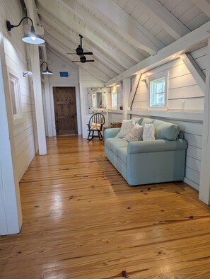 A spacious room with wooden flooring, a light blue sofa, a rocking chair, and a ceiling fan. - Canoe Club Loft - Dock slip available (Daufuskie Island)