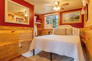 A wooden cabin bedroom with a bed, a ceiling fan, a window with a view of trees, and a painting on the wall. - Cody Bunkhouse (Cody)