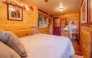 Une pièce de cabane en bois comprenant un lit, une fenêtre avec des rideaux, un miroir et une porte donnant sur une autre pièce.