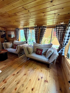 Ein gemütliches Blockhaus-Interieur mit Holzböden, einer Couch mit Kissen, einem kleinen Tisch und einem Fenster mit Vorhängen.