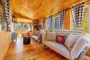 Ein gemütliches Blockhauss-Interieur mit Holzbalkendecke, einer Couch, einem Tisch und Blick ins Freie.