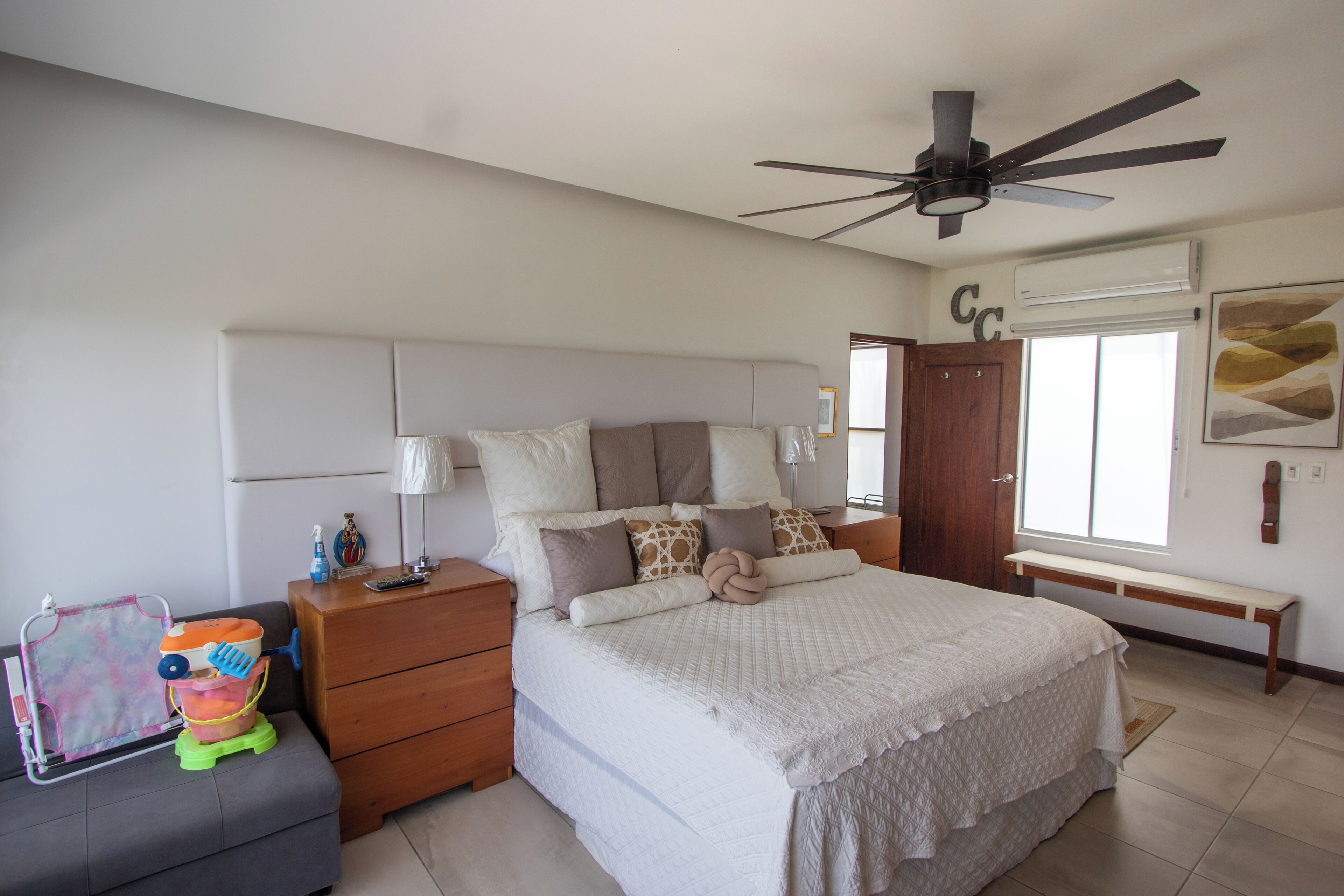 Une chambre à coucher comprenant un lit, une table de chevet, une commode, un ventilateur de plafond, une fenêtre et un banc.