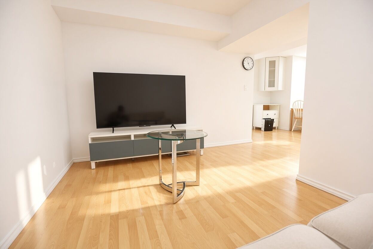 A modern living room with a flat-screen TV, a glass-top coffee table, and a kitchen area in the background.
