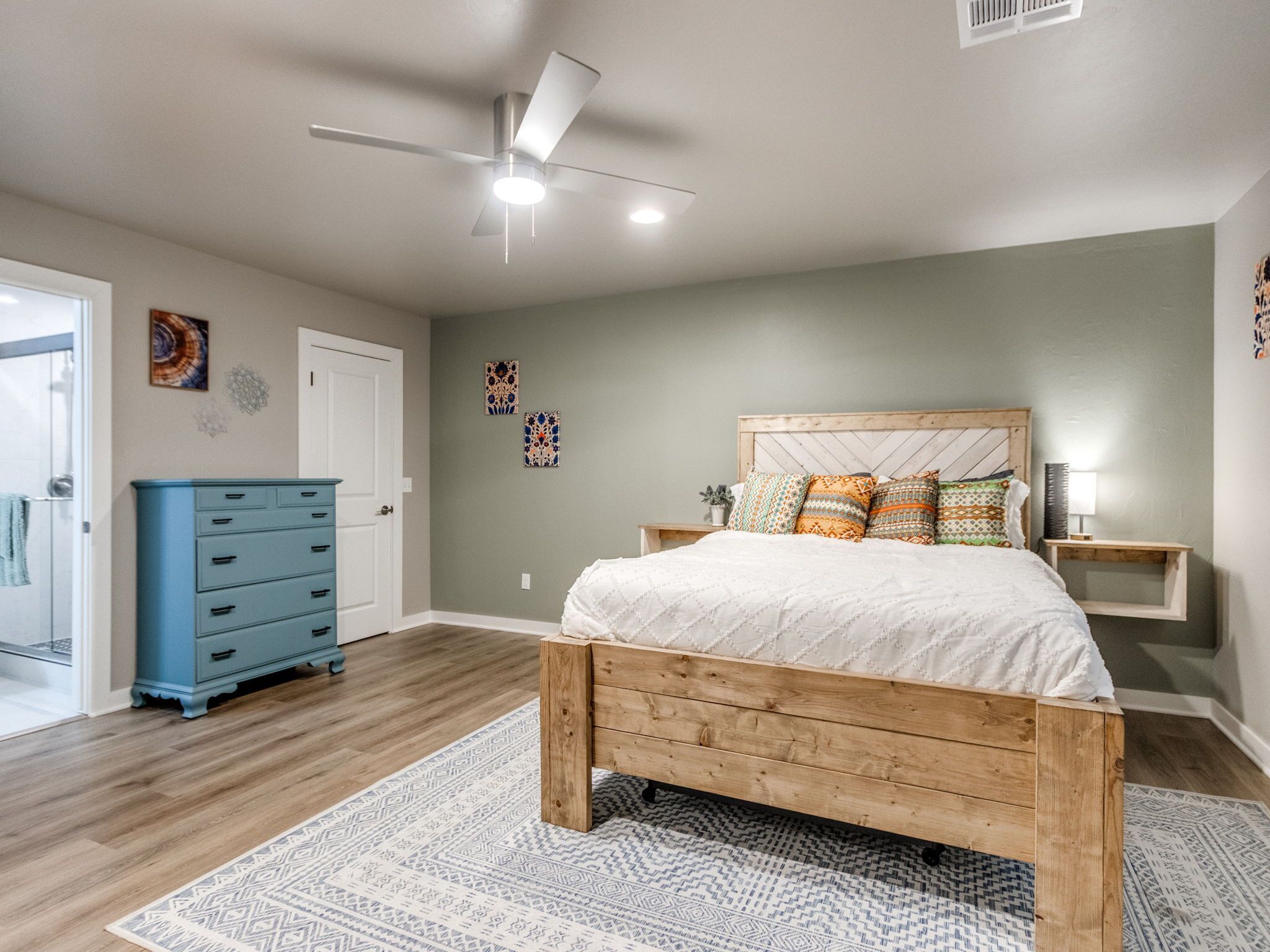 Un dormitorio con una cama de madera, un ventilador de techo, un aparador azul y alfombras estampadas.