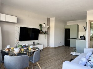 A modern living room with a dining area, a flat-screen TV mounted on the wall, a kitchen area, and a sofa. - Flower Beach Helgueras 2 - 6092 (Noja)