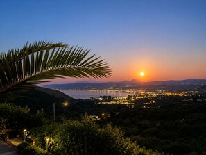 A sunset view over a coastal city with illuminated buildings and a palm tree in the foreground. - Oceana Suites Villas Deluxe Private Pool Studio (Apokoronas)
