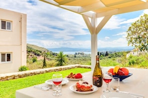 Tisch im Freien mit einer Flasche, Gläsern und Obst auf einer Terrasse mit Blick auf Hügel und ein Gebäude.