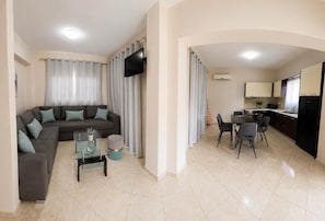 Ein Wohnzimmer mit Sofa, Couchtisch und Fernseher. Es gibt einen Essbereich mit Tisch und Stühlen. Die Küche ist sichtbar, mit Schränken und Geräten.