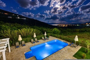 A swimming pool with lounge chairs and umbrellas, surrounded by greenery and a view of a distant cityscape at night. - Oceana Suites Villas - Spacious Family Suite (Apokoronas)