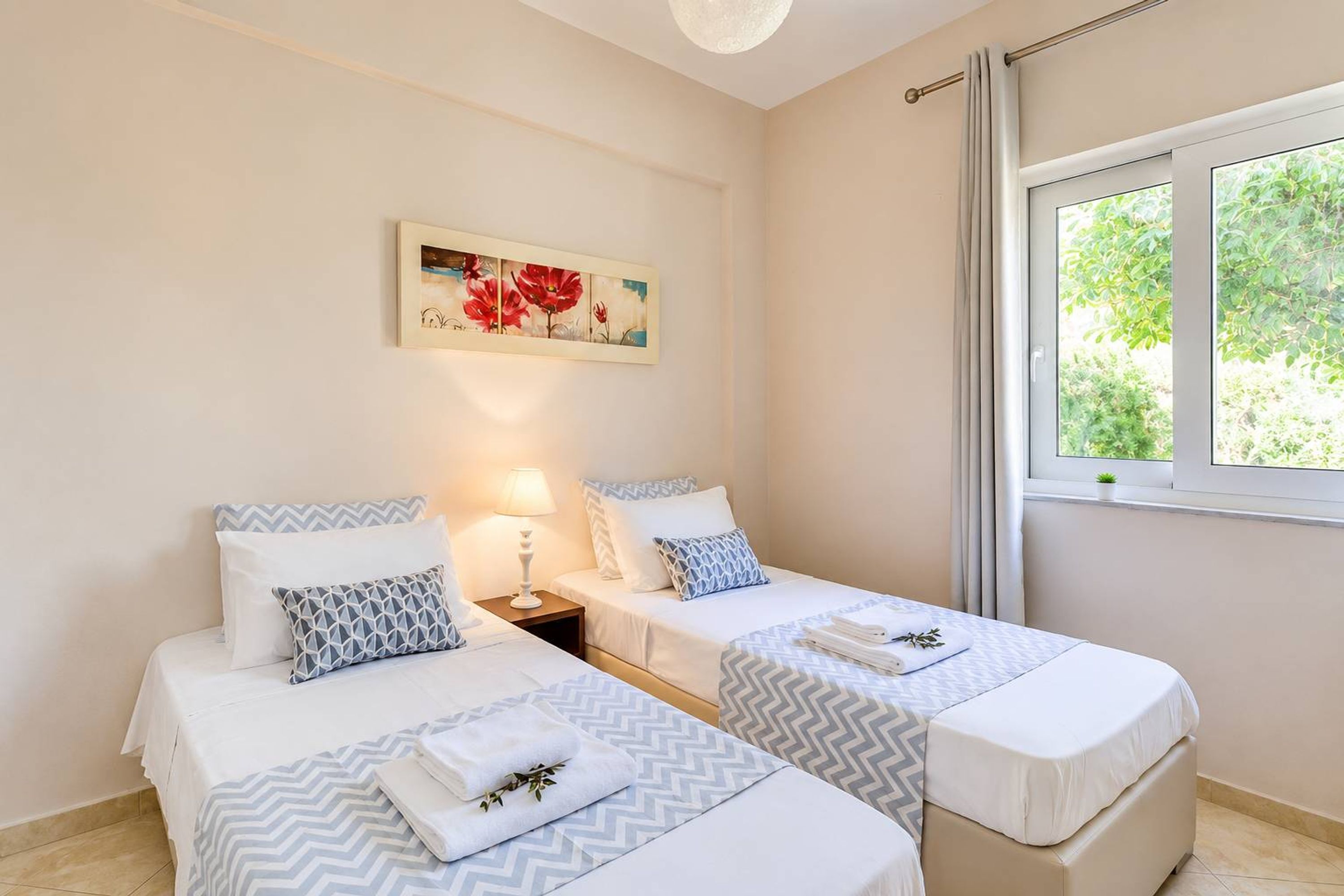 A hotel room with two beds, a nightstand, a lamp, a window with curtains, and a painting on the wall.