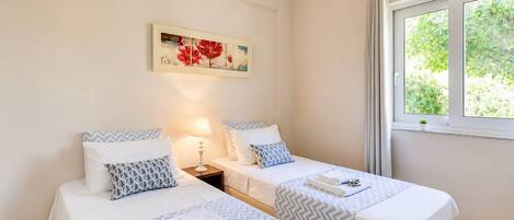 A hotel room with two beds, a nightstand, a lamp, a window with curtains, and a painting on the wall.