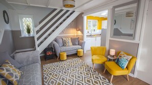 Un espace de vie compact comprenant un canapé, des fauteuils, un tapis à motifs, un escalier et un coin cuisine.