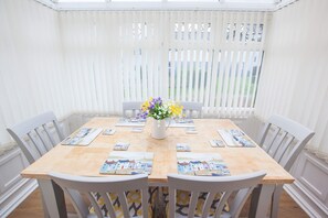 A dining area with a wooden table, white chairs, a vase with flowers, and a view through white blinds. - Poachers Cottage Old Tom Cottage Sleeps 12 Dog Friendly Pool Tennis Family Facilities (Tenby)