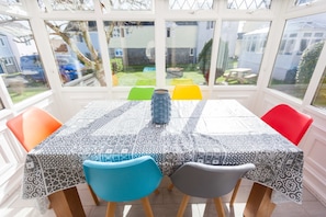 A conservatory with a table covered in a patterned tablecloth, surrounded by colorful chairs.