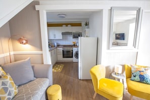 A compact living space with a kitchenette, a sofa, a yellow armchair, and a small table with a lamp.