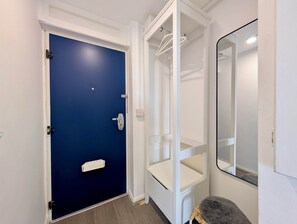 Ein Zimmer mit blauer Tür, weißen Wänden, einem weißen Kleiderschrank mit Kleiderstange und einem Spiegel an der Wand.