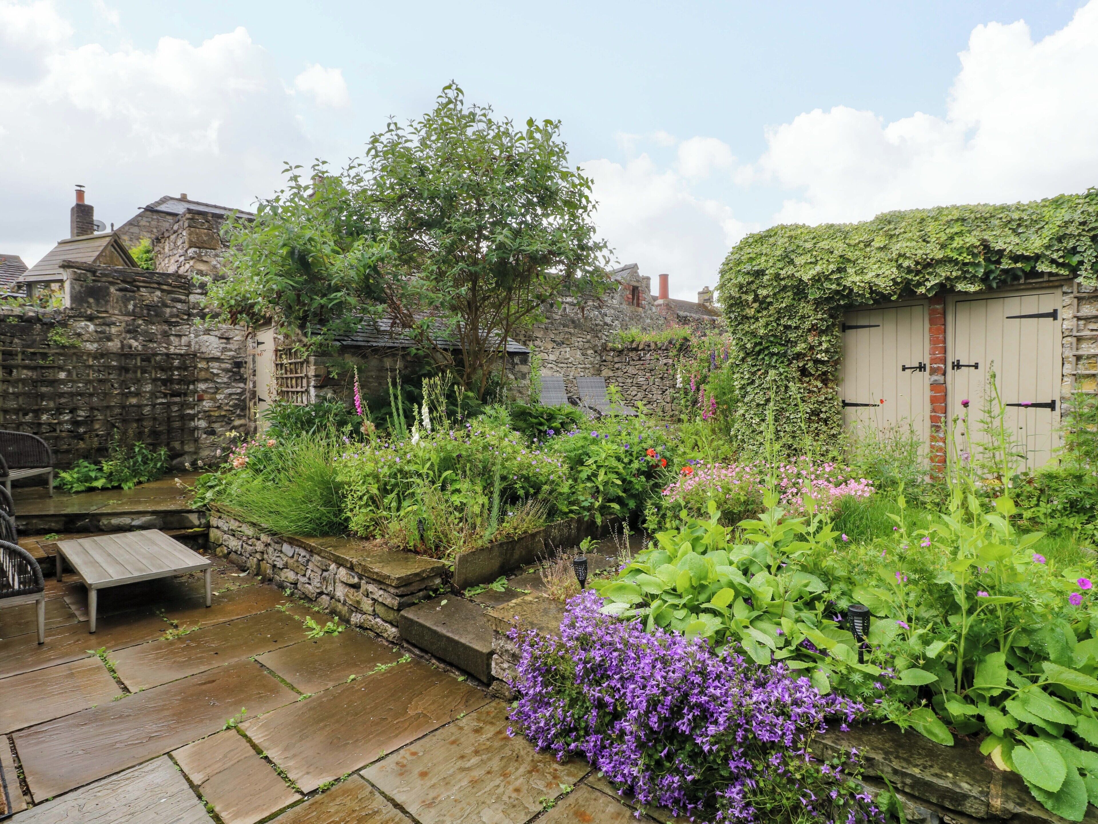 Un patio de piedra con un banco de madera, un muro de piedra y un jardín con diversas plantas y flores.