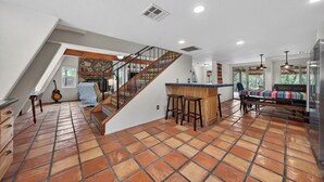 Ein geräumiger Wohnbereich mit Fliesenboden, einer Treppe mit dekorativer Geländer und einer Küchenzeile mit Bar, sowie eine Sitzecke mit farbenfroher Couch.