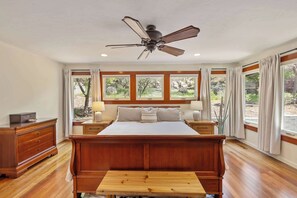 Ein Schlafzimmer mit einem hölzernen Bett, einem Deckenventilator, einem Nachttisch mit Lampe und großen Fenstern mit Vorhängen.