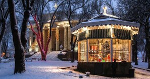 Ein mit Schnee bedeckter Pavillon mit einem Weihnachtsbaum im Inneren, umgeben von kahlen Bäumen und einem Gebäude mit Säulen im Hintergrund.