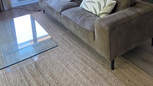 Una sala de estar con sofá, una mesa de centro de vidrio, lámpara, mesa de comedor con sillas y estantería.