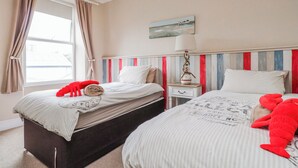 A bedroom with two beds, a window with curtains, a lamp, and a painting on the wall. - The View (Criccieth)