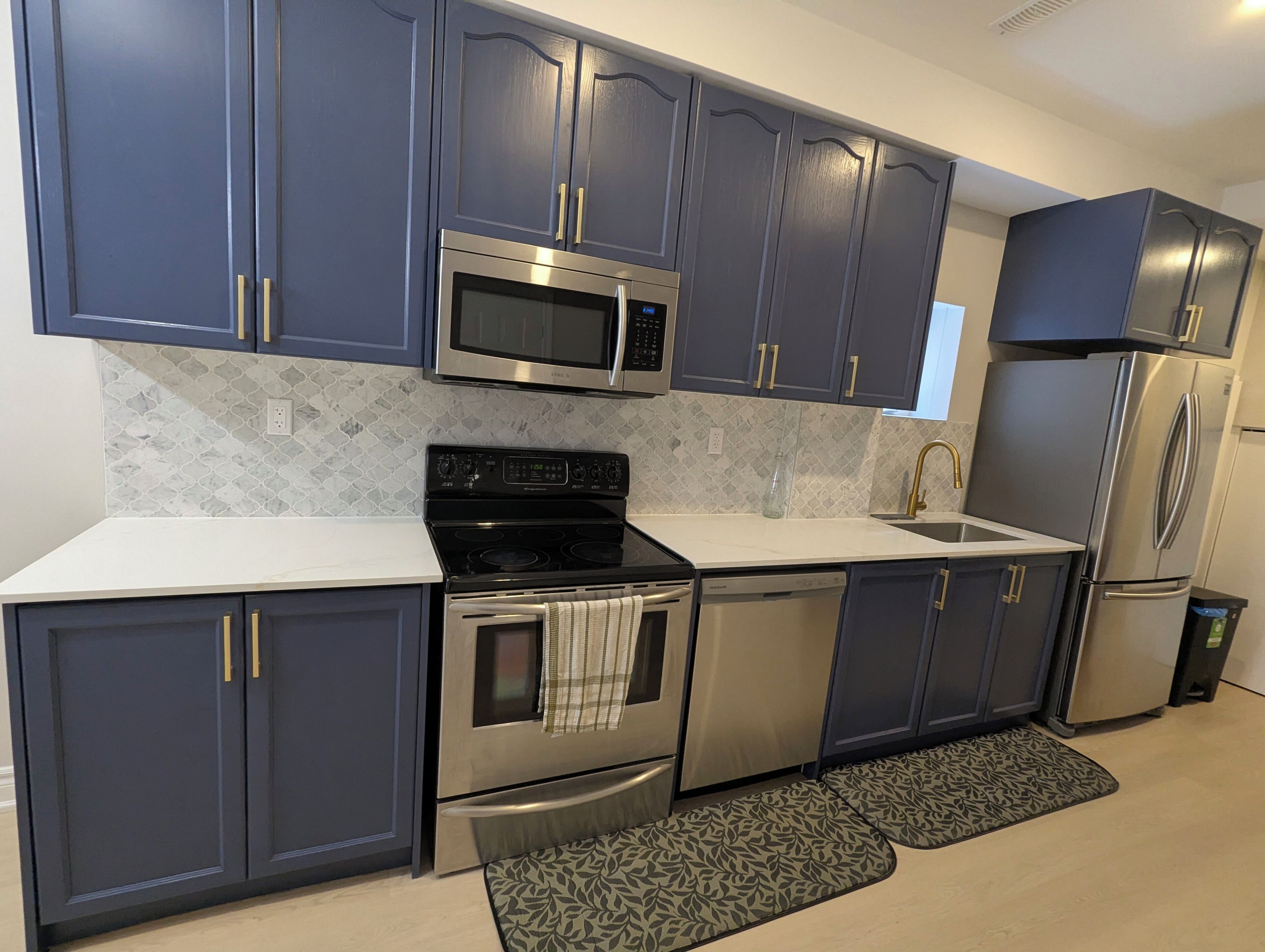 A modern kitchen with dark blue cabinets, stainless steel appliances, a white countertop, and a patterned rug.