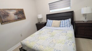 Een slaapkamer met een bed, een nachtkastje, een lamp, een raam met jaloezieën en een schilderij aan de muur.