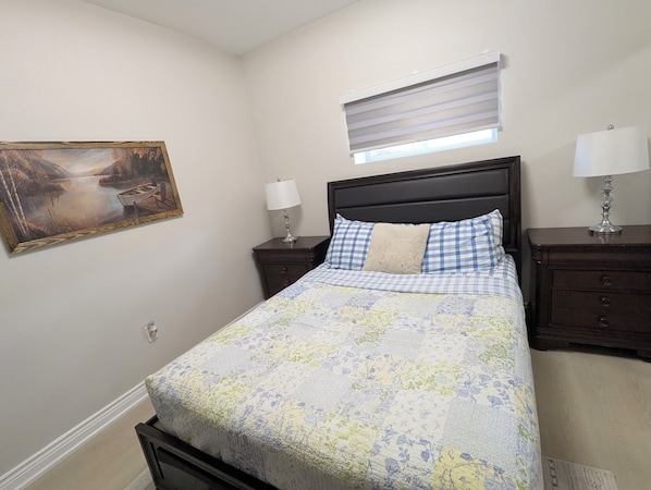 A bedroom with a bed, a nightstand, a lamp, a window with blinds, and a painting on the wall. - Modern Designer Suite — Minutes to Thermëa Spa & Highways 407, 401 and GO (Whitby)