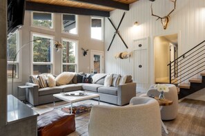 A modern living room with a gray sectional sofa, a coffee table, and a fireplace. There are large windows and a staircase leading to another level. - West Vail Home - Hot Tub, Fireplace & Steps to Bus (Vail)
