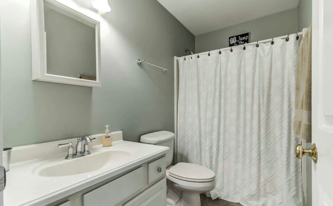 Un baño con lavamanos, espejo e inodoro blancos, cortina de ducha y un cartel sobre la ducha.