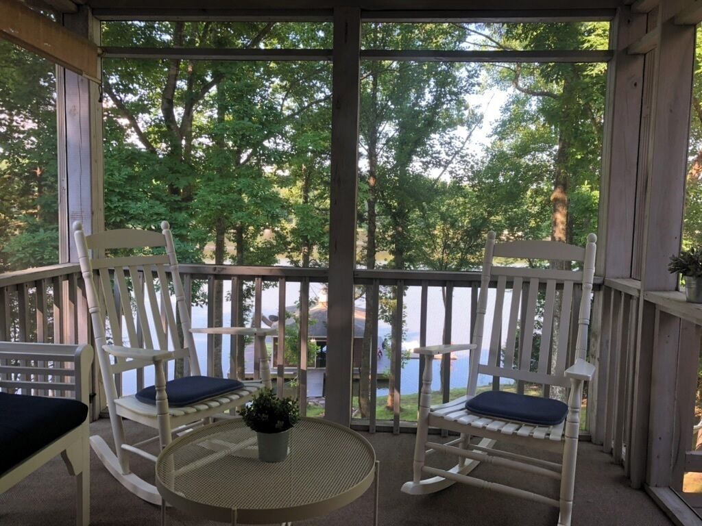 A covered porch with two rocking chairs and a table, overlooking a body of water and greenery.