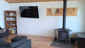 Una sala de estar con piso de madera, un sofá, un televisor montado en la pared, una estantería y un estufa de leña.