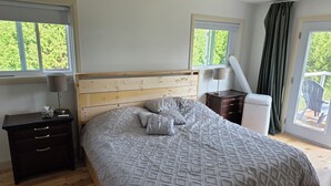 Un dormitorio con cabecera de madera, cama con ropa de cama estampada, mesita de noche con lámpara, cómoda y ventana con vista a follaje.