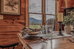 A wooden interior with a dining table set for two, a candle, and a view of mountains through a window. - Summit Views | Hot Tub, Sauna, Fire Pit & Game Room, Near Gatlinburg (Gatlinburg)