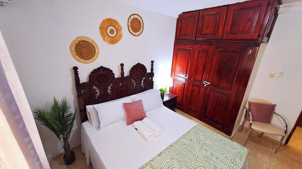 Une chambre à coucher avec une tête de lit en bois, un lit recouvert d’une literie blanche, un plaid à motifs verts, une armoire en bois et une petite chaise.