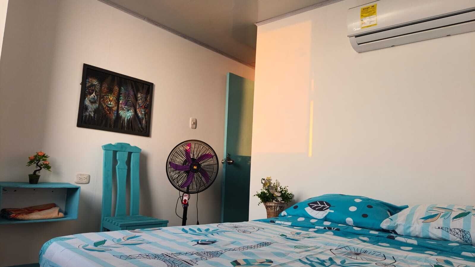 A bedroom with a bed, a fan, a wall-mounted air conditioner, a door, and a shelf with a plant.