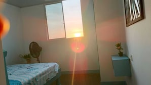 Ein Zimmer mit einem Bett, einem Standventilator, einem Fenster und einer kleinen Pflanze auf einem Regal.