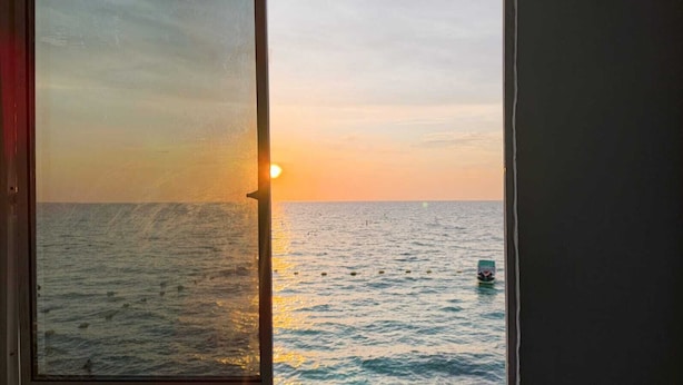 Blick durch ein Fenster auf einen Sonnenuntergang über dem Meer mit einem Schiff in der Ferne.