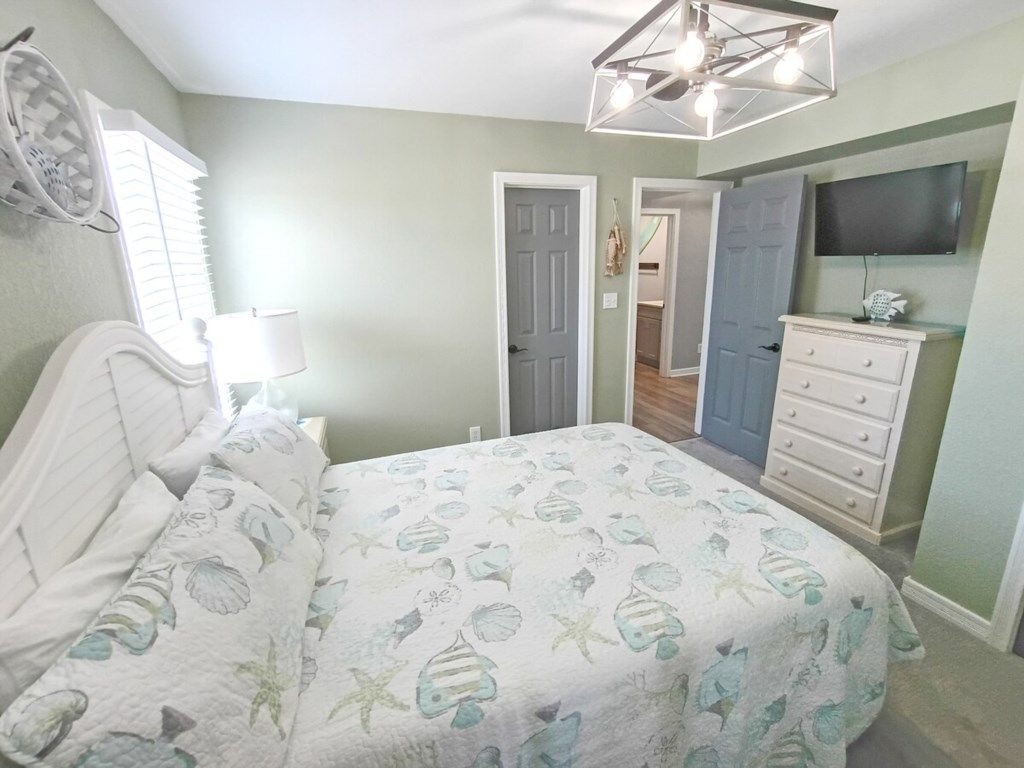 Une chambre à coucher comprenant un lit, une commode, un téléviseur et une porte donnant sur une autre pièce.