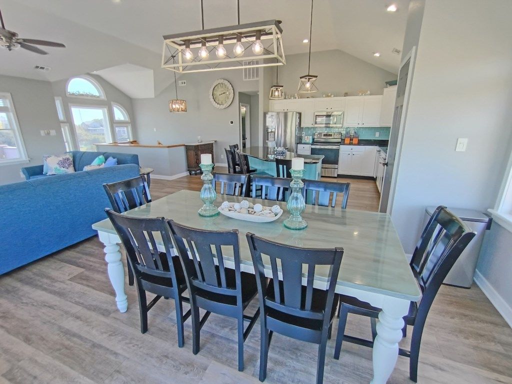 Un grand espace à manger avec une grande table et des chaises, une cuisine en arrière-plan et un ventilateur de plafond.