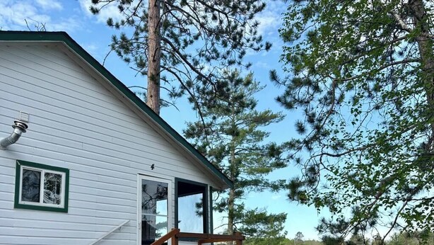 Une cabane blanche avec un escalier en bois menant à une terrasse, entourée d’arbres et bordée par un plan d’eau.
