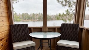 Une pièce avec deux fauteuils en osier et une table en verre, un plancher en bois et une grande fenêtre donnant sur un lac et des arbres.